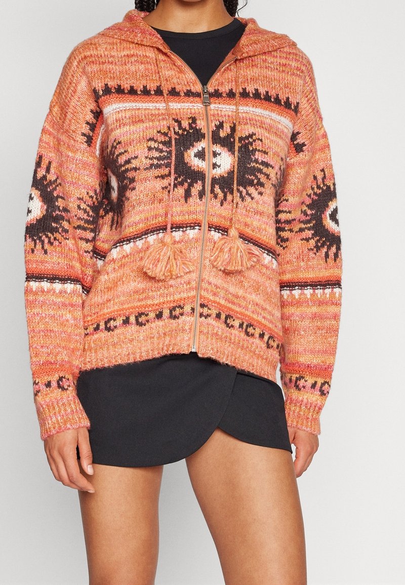 Personne portant un sweat à capuche en maille orange zippé avec des motifs soleils noir et blanc, des pompons sur les cordons de serrage, et une jupe asymétrique noire.