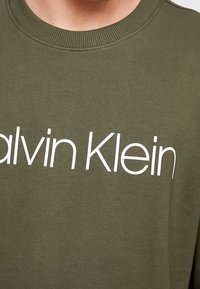 Felpa verde oliva in cotone con colletto tondo e il logo bianco "Calvin Klein" ben visibile sulla parte anteriore.