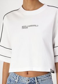 Balta trumpa marškinėliai su apvaliu iškirpimu, papuošti juodu apvadu aplink rankoves ir Karl Lagerfeld Jeans logotipu siuvinėtu priekyje.