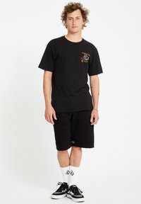 T-shirt noir avec logo sur le devant, associé à un short noir. Le modèle porte des baskets noires et des chaussettes blanches avec logo, se tenant devant un fond blanc.