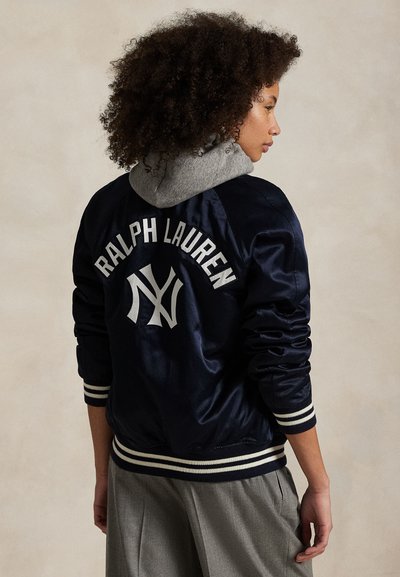 Göndör hajú személy, aki sötétkék szatén dzsekit visel, amelyen a "Ralph Lauren" és "NY" logók vannak a háton, szürke nadrággal és szürke kapucnival párosítva.