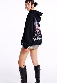 Sort oversize hættetrøje med grafik på ryggen. Kombineret med skinnende sølvshorts og knæhøje sorte støvler med spænder.
