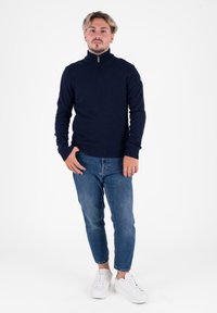 Dunkelblauer Zip-Neck-Pullover aus strukturiertem Strick, kombiniert mit hellblauen Jeans und weißen Sneakers. Der Pullover hat gerippte Bündchen und einen gerippten Saum.