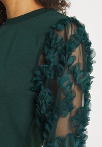 Green long-sleeve top with sheer mesh sleeves adorned with textured floral appliques on the arm.