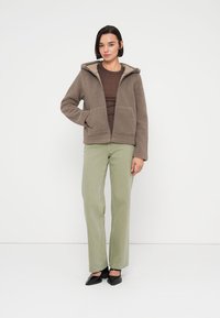 Καφέ φλις μπουφάν με κουκούλα, φερμουάρ μπροστά και δύο τσέπες, σε συνδυασμό με ελαιί πράσινο παντελόνι με ίσια γραμμή και μαύρα παπούτσια.