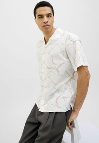Camisa de manga corta de tela blanca con un patrón abstracto en rosa y beige. Incluye un cuello con muesca y cierre de botones. Se lleva con pantalones gris.