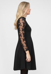Vestido negro con corpiño ajustado y falda acampanada. Mangas largas transparentes con detalles de encaje floral negro. Tela suave en el cuerpo principal.