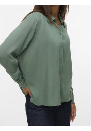 Vrouw die een loszittend, lang mouw button-down overhemd in saliegroen draagt, met donkerblauwe jeans, staand met één hand in de zak.