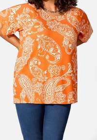 Haut orange avec motifs paisley et floraux blancs, manches courtes et col rond, associé à un pantalon en denim bleu foncé.