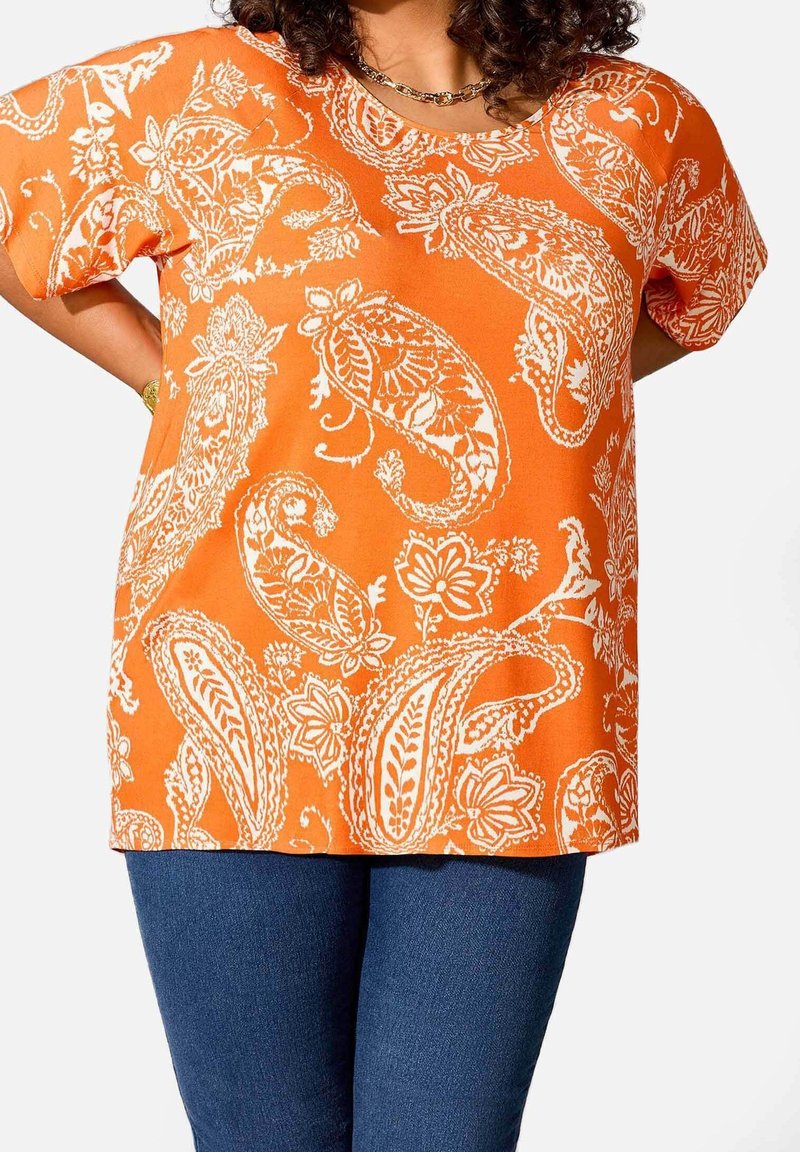 Haut orange avec motifs paisley et floraux blancs, manches courtes et col rond, associé à un pantalon en denim bleu foncé.