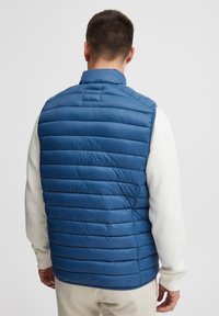 Mann in einer blauen, gesteppten ärmellosen Weste über einem weißen Langarmhemd, mit dem Rücken zu einer schlichten weißen Wand stehend.