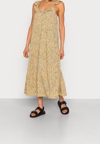 Robe fleurie jaune avec bretelles à volants ; longueur mi-mollet, tissu léger ; associée à des sandales noires épaisses. Motif texturé sur toute la robe.