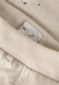 Beigefarbenes Gewebe mit einem gerippten elastischen Bund, der mit einem Knopfverschluss versehen ist. Ein leichtes Muster ist im unteren Bereich sichtbar, das subtile Designdetails zeigt.