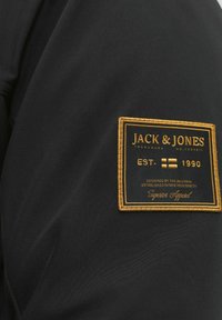 Černá tkanina s obdélníkovou nášivkou prošívanou zlatou nití, na které je nápis "JACK & JONES," "EST. 1990" a další brandingové detaily.