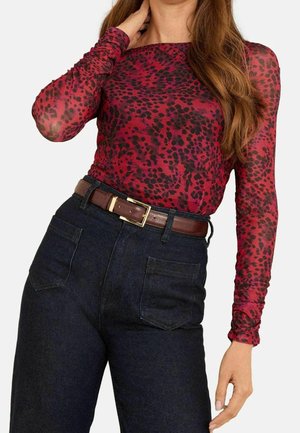 Vrouw met een lange mouwen, rood-zwart luipaardprint top die is ingestopt in een hoog getailleerde donkere spijkerbroek met een bruine riem.
