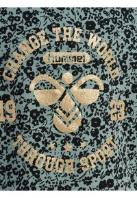 Grün gemustertes Stoff mit goldglänzenden Texten und Akzenten. Zu den Merkmalen gehören eine zentrale Biene Grafik und der Text "ÄNDERE DIE WELT DURCH SPORT".