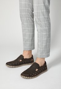 Braune Slipper mit durchbrochener Oberseite und Ausschnittdetails, ausgestattet mit einer strukturierten Sohle mit kontrastierenden beigen Akzenten. Getragen zu grauen Karohosen.