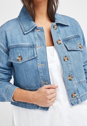 Femme portant une veste en jean bleu clair avec de gros boutons marron sur une robe blanche à œillets, main ajustant le devant de la veste.