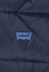 Veste matelassée bleu marine avec un design matelassé, arborant un patch logo Levi's en caoutchouc bleu sur la poitrine. Texture lisse et brillante.