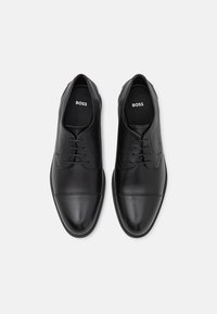 Chaussures habillées en cuir noir avec une texture lisse, bout pointu, cinq œillets et un discret motif cousu sur les côtés. L'intérieur présente le logo de la marque.