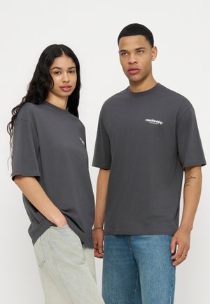 Δύο νεαροί ενήλικες στέκονται δίπλα-δίπλα φορώντας υπερμεγέθη σκούρα γκρι μπλουζάκια με το κείμενο «overthinking» στο στήθος, συνδυασμένα με ανοιχτόχρωμα τζιν.