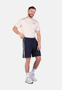 Dunkelblaue Shorts mit weißen Seitenstreifen und Logo, kombiniert mit einem hellbeigen T-Shirt. Weiße Socken und graue Sneakers runden das Outfit ab.