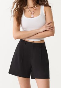 Černé elegantní šortky s hladkou strukturou, s předními zřaseními a pasem. V kombinaci s bílým žebrovaným tílkem a náhrdelníkem.
