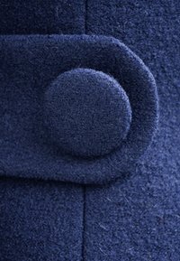 Granatowa wełniana tkanina o fakturowanej powierzchni. Posiada zaokrąglony guzik oraz złożony klapę po prawej stronie, podkreślając głębię materiału.