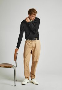 Camisa negra de manga larga, pantalones de lona beige con dos bolsillos frontales, cinturón negro y zapatillas blancas. De pie junto a una silla.