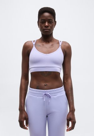 Sportos személy áll, előre nézve, világos lila sportmelltartót és hozzá illő, magas derekú, zsinórral állítható melegítőnadrágot visel.