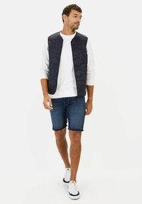 Marineblaue gesteppte Weste mit hohem Kragen, kombiniert mit einem weißen Sweatshirt und Jeansshorts. Weiße Turnschuhe mit marineblauen Akzenten runden das Outfit ab.