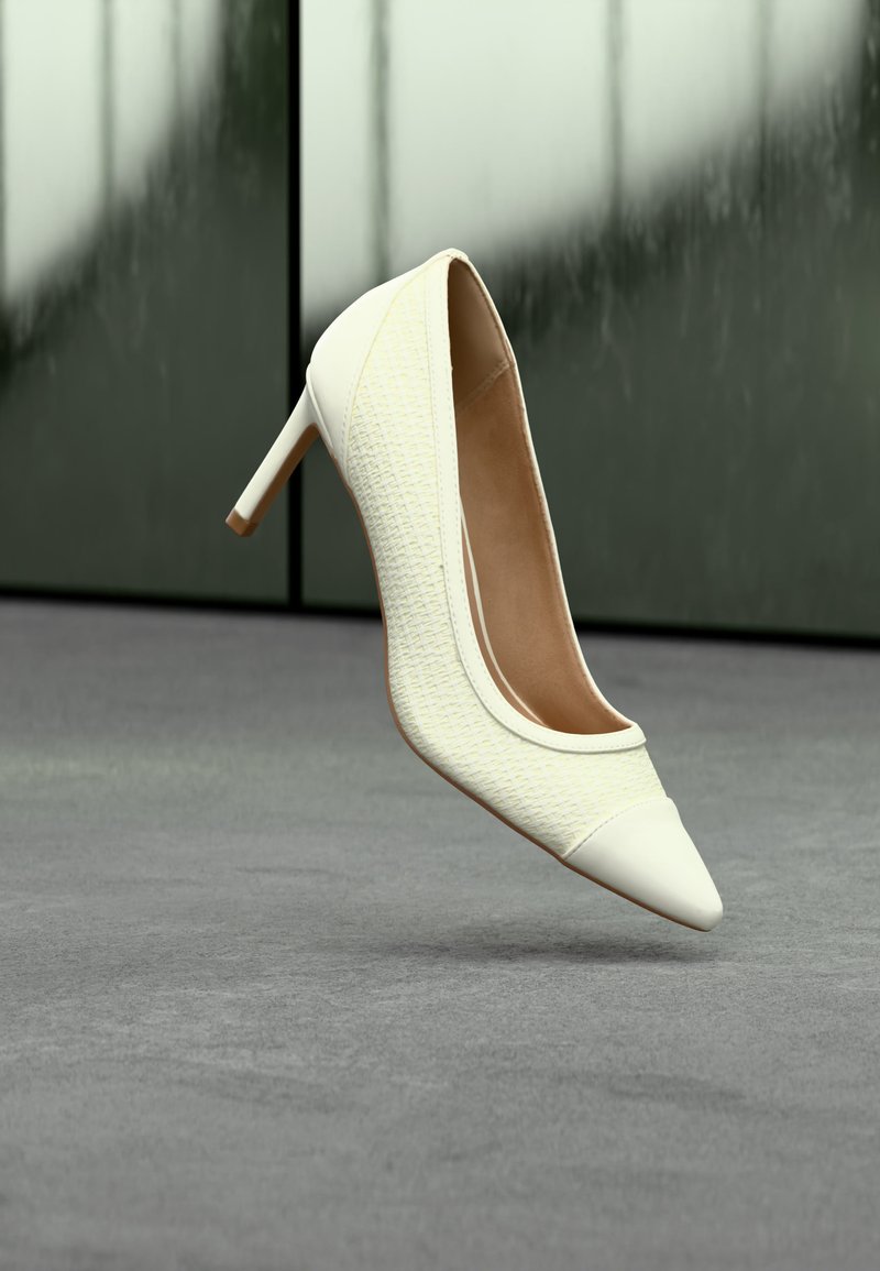 Zapato blanco de tacón alto con tela texturizada y punta afilada, flotando sobre un suelo alfombrado gris con fondo de cristal desenfocado.