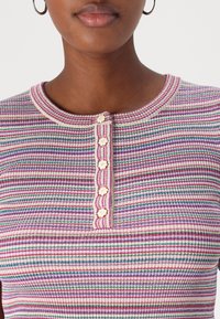 Personne portant un t-shirt à rayures roses, blanches et vertes, avec cinq boutons en forme de fleur sur le devant et des boucles d'oreilles en or.