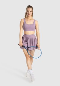 Top corto sportivo viola e gonna plissettata, abbinati a una racchetta da tennis e sneaker bianche. Finitura opaca, texture liscia.