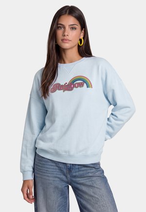 Donna con lunghi capelli scuri che indossa orecchini a cerchio gialli, una felpa azzurro chiaro con "Rainbow" e una grafica arcobaleno, e jeans blu.