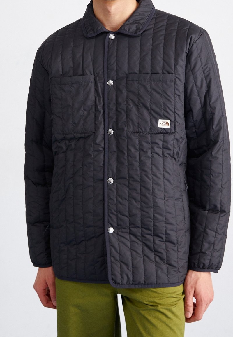 Quiltet sort jakke med knaplukning fortil, to brystlommer og The North Face-logo på venstre lomme. Enkel krave-design.