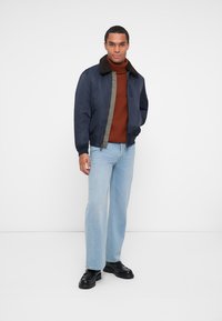 Veste bombardier marine avec un col en fausse fourrure, doublure matelassée et fermeture éclair, associée à un col roulé rouille et un jean bleu clair.