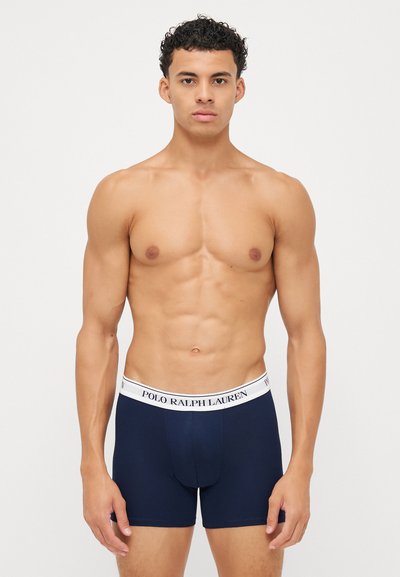 Mornarske bokserice v navy barvi z belim elastičnim pasom, na katerem je napis "POLO RALPH LAUREN". Gladka tkanina, prilegajoča zasnova, dolžina do sredine stegna.