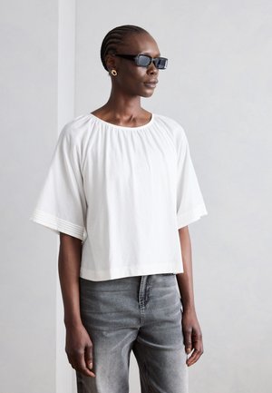 Kvinde med cornrow-frisure iført sorte rektangulære solbriller, gyldne hoop-øreringe, løs hvid top og falmede grå jeans, står sideværts.