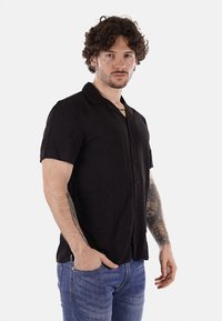 Camicetta nera a maniche corte con chiusura a bottoni, colletto piatto e texture liscia, abbinata a jeans blu.