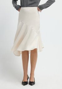 Vrouw in een lichtbeige asymmetrische midi-rok, grijze lange mouwen top en zwarte puntige hakken, staande met haar handen op haar heupen.