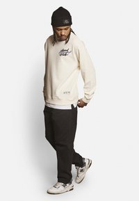 Sudadera crema con texto gráfico, acentos negros y puños de canalé, combinada con pantalones negros y zapatillas blancas con detalles grises.