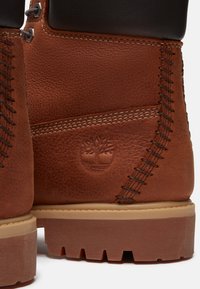 Tacones de botas de cuero marrón con detalles de costuras visibles, suelas texturizadas y logotipo de árbol en relieve en el panel lateral.