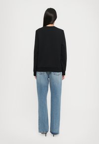 Donna con lunghi capelli lisci, che indossa un felpa nera e jeans dritti blu chiaro, in piedi di spalle contro uno sfondo bianco.