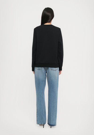 Femme aux longs cheveux raides portant un sweat-shirt noir et un jean droit bleu clair, se tenant de dos devant un fond blanc.