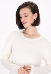 Könnyű krémszínű pulóver kerek nyakkivágással, hosszú ujjakkal és bordázott mandzsettákkal. Sima textúra minimális varrási részletekkel.