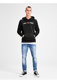 Sort hættetrøje med hvidt logo, frontlomme, snore. Lyseblå slidte jeans, afslappede sorte og hvide sneakers. Enkel design.