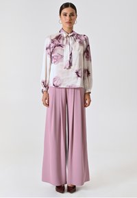 Blusa a maniche lunghe bianca con motivo floreale viola, abbinata a pantaloni rosa a vita alta e gamba larga. Modello con scarpe color bordeaux.