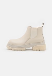 Botas de cuero beige hasta el tobillo con una suela blanca gruesa, paneles laterales elásticos y una lengüeta en la parte posterior; textura suave y diseño moderno.