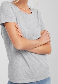 Szara koszulka z krótkim rękawem i okrągłym dekoltem, wykonana z miękkiego materiału. Koszulka ma luźny krój i gładką strukturę, noszona z założonymi na siebie rękami.